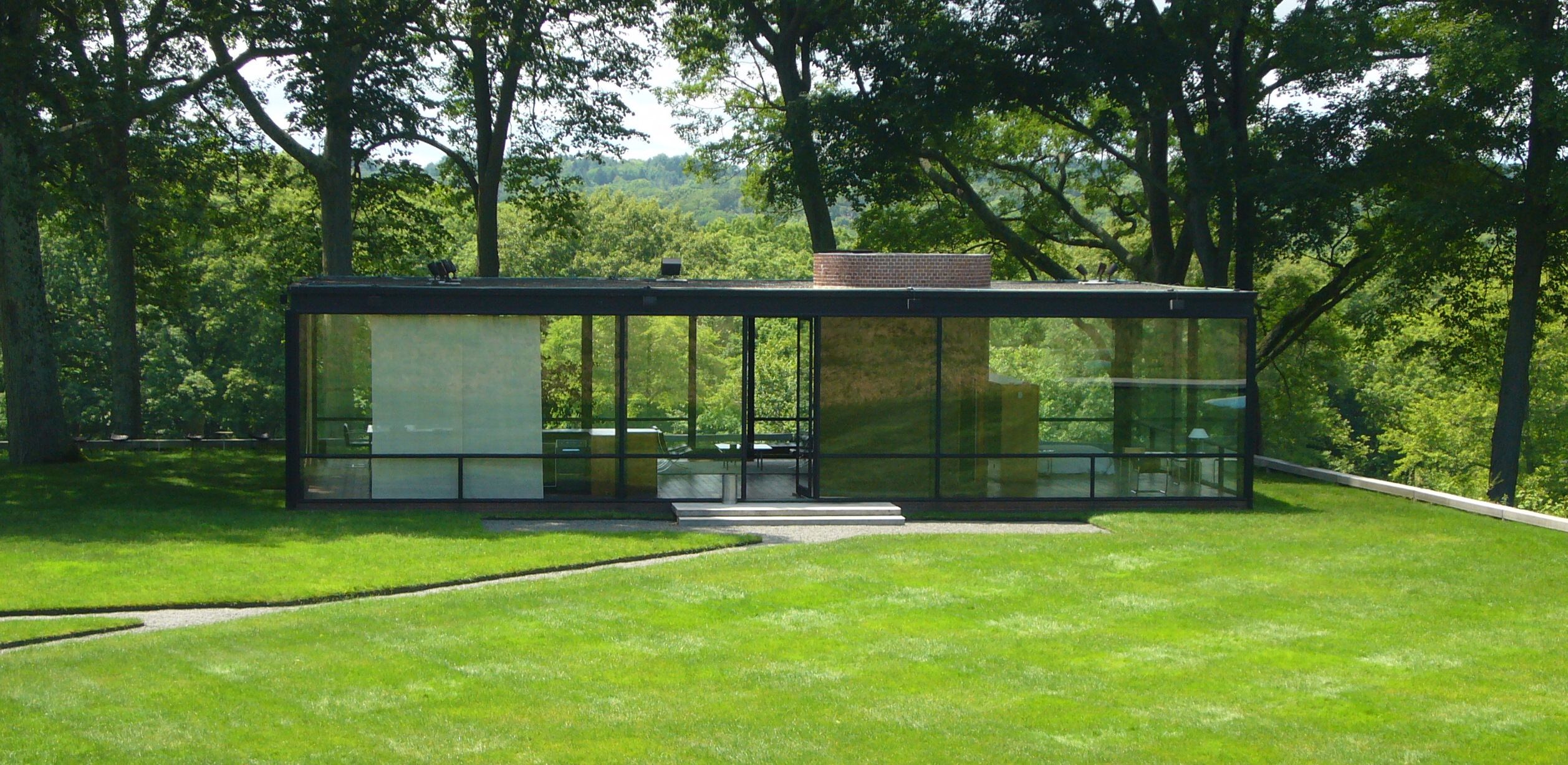 blog arquitectos 24 Vista frontal de la Glass House de Philip Johnson, ejemplo icónico de transparencia arquitectónica en medio de un entorno natural arbolado.