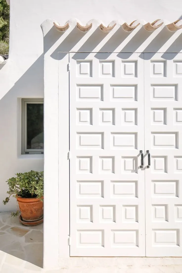 Diferencias entre la arquitectura vernácula de viviendas en Alicante y Murcia 5 Puerta doble blanca con molduras geométricas en fachada encalada, con maceta de terracota y tejas curvas, ejemplo de arquitectura vernácula mediterránea.